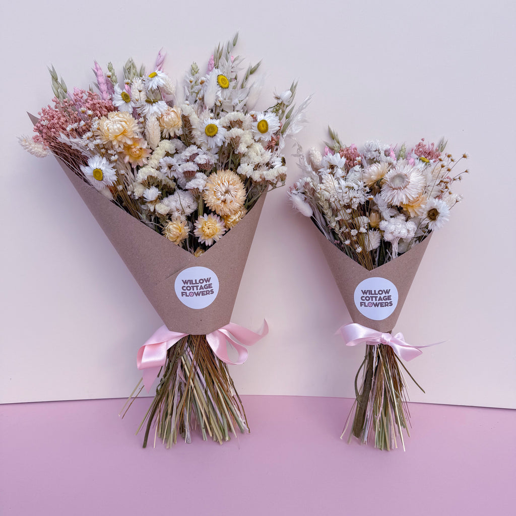 Two small dried flower bouquets wrapped in brown paper cones with pink ribbons, displayed against a split white and light pink background. Each bouquet has a round white label reading 'WILLOW COTTAGE FLOWERS'.