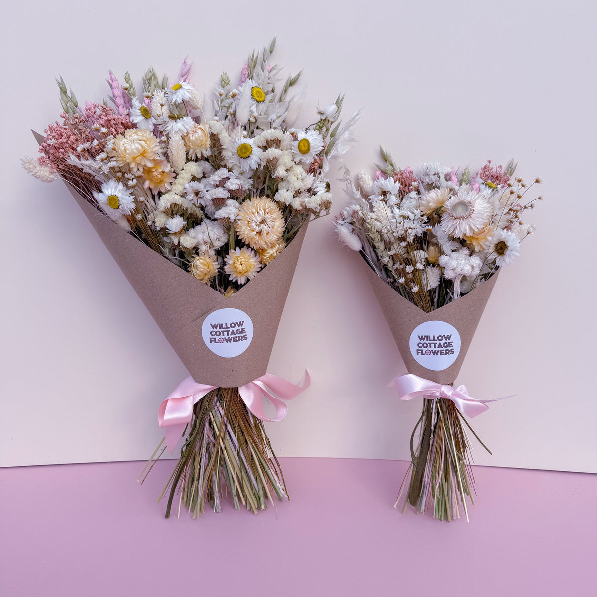 Two small dried flower bouquets wrapped in brown paper cones with pink ribbons, displayed against a split white and light pink background. Each bouquet has a round white label reading 'WILLOW COTTAGE FLOWERS'.