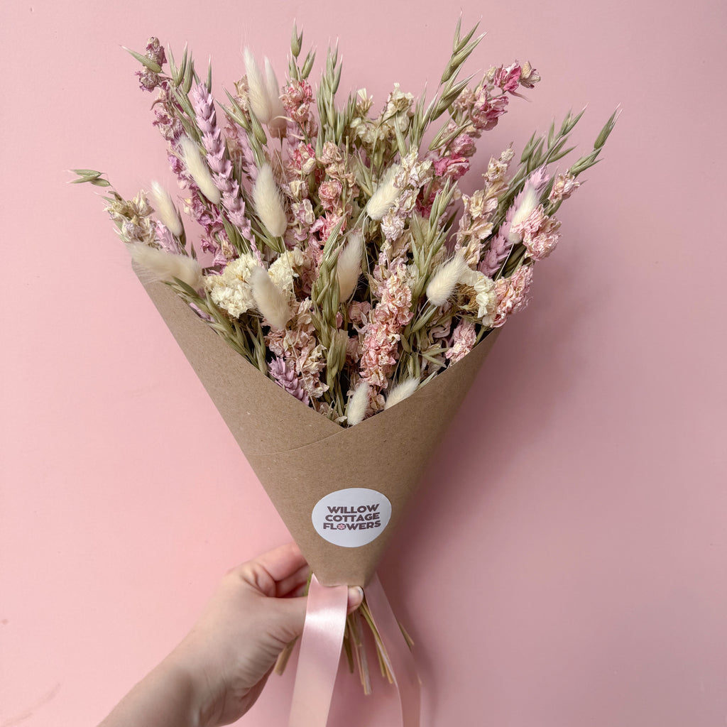 A hand holding a bouquet of dried flowers wrapped in brown kraft paper with a pink ribbon. The bouquet features soft pink, white, and beige dried blossoms.