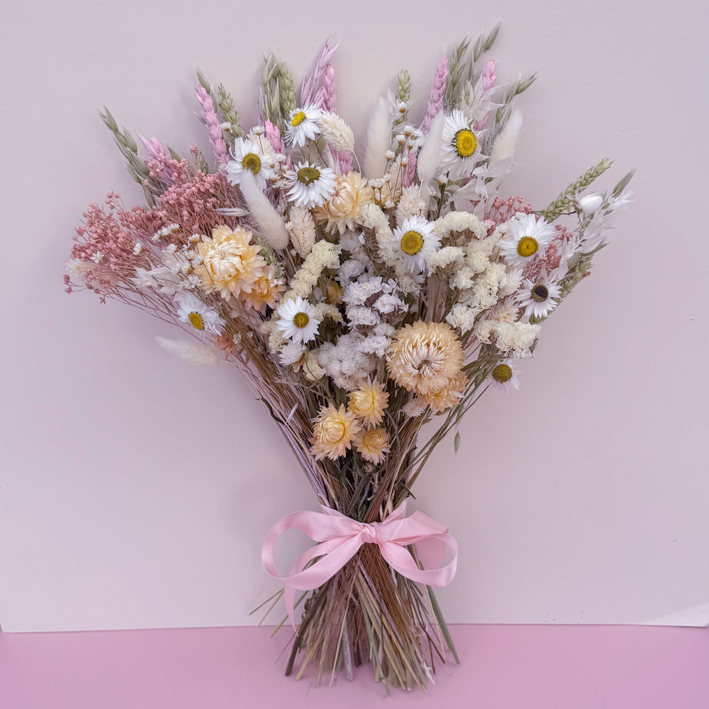 A dried flower bouquet featuring white daisies, pink and cream blossoms, wheat stems, and fluffy bunny tails, tied with a light pink ribbon against a soft pink background.