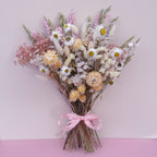 A dried flower bouquet featuring white daisies, pink and cream blossoms, wheat stems, and fluffy bunny tails, tied with a light pink ribbon against a soft pink background.