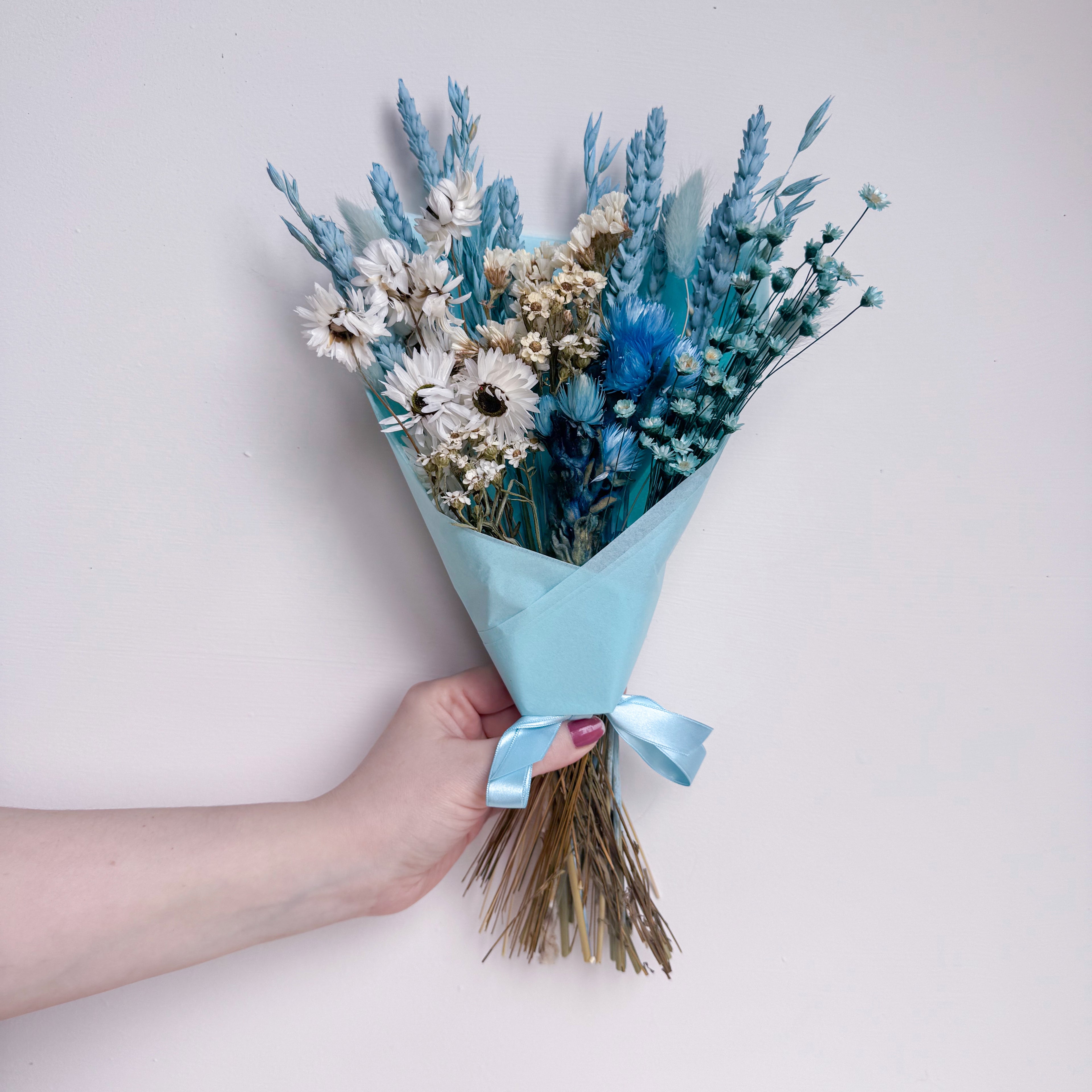 Bouquet of blue and white flowers wrapped in light blue paper held by a hand on a light background