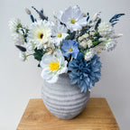 Gray textured vase with a bouquet of white, blue, and yellow artificial flowers on a wooden surface.