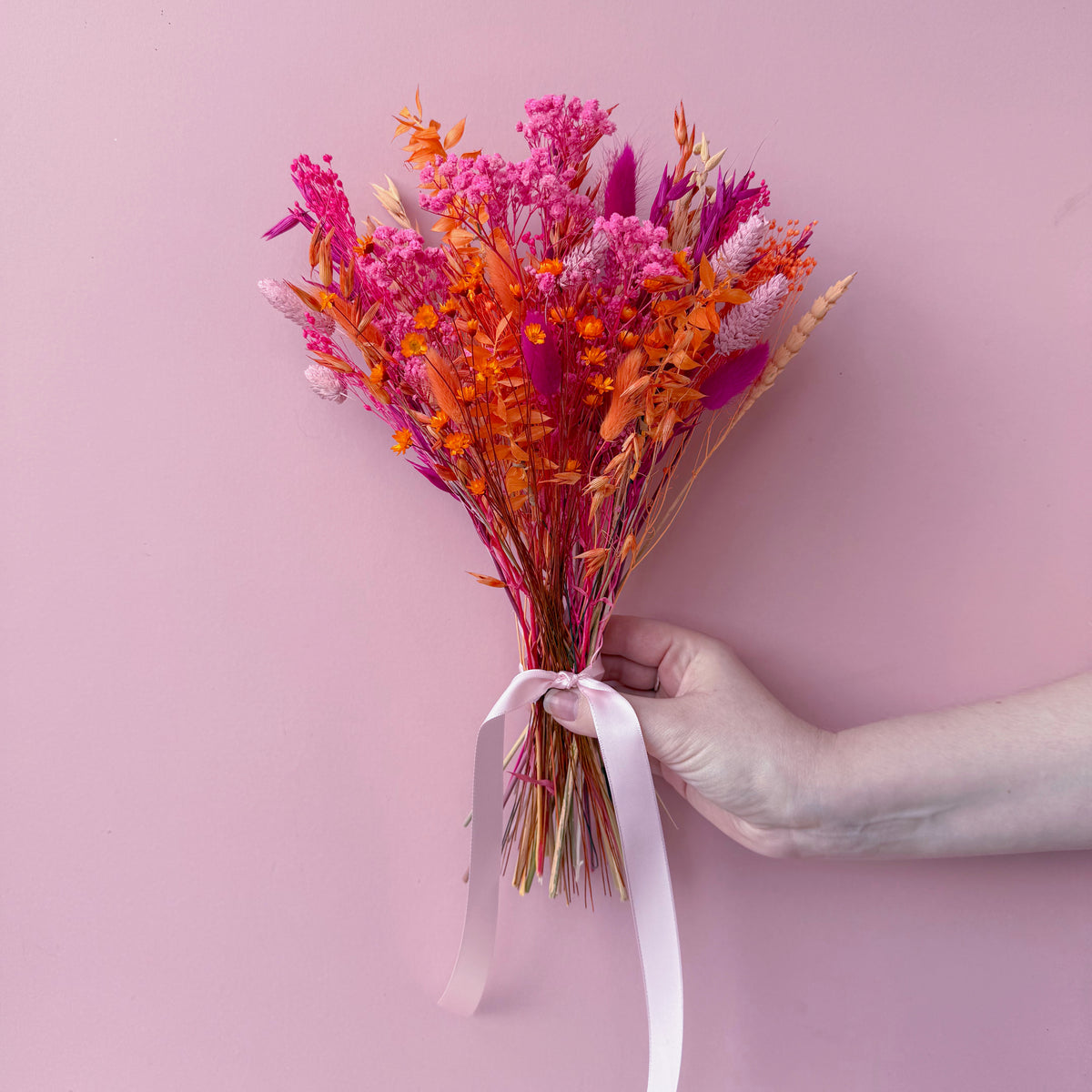 A hand holding a small bouquet of dried flowers in shades of pink, orange, and red tied with a pale pink ribbon, set against a solid pink background.