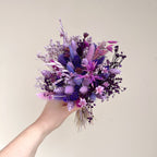 A hand holding a dried flower bouquet in shades of purple, lavender, and pink against a plain beige background.
