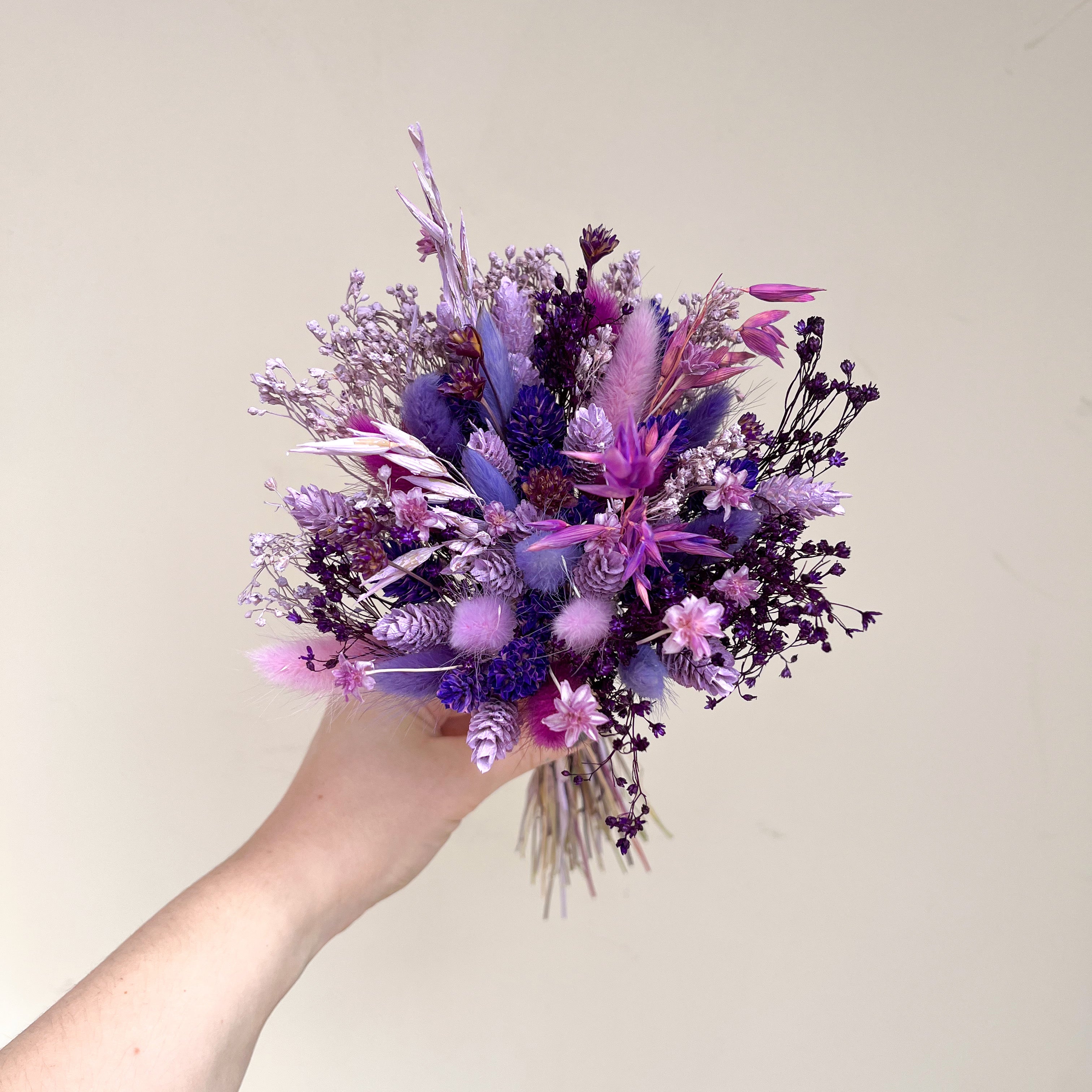 A hand holding a dried flower bouquet in shades of purple, lavender, and pink against a plain beige background.