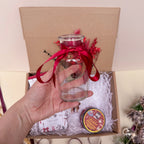 Clear glass vase with red ribbon held over a gift box with decorative items.
