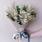 A green dried flower bouquet wrapped in brown kraft paper with a white round sticker reading 'WILLOW COTTAGE FLOWERS' and tied with a dark green and white ribbon, held by a person's hand against a pale pink background.