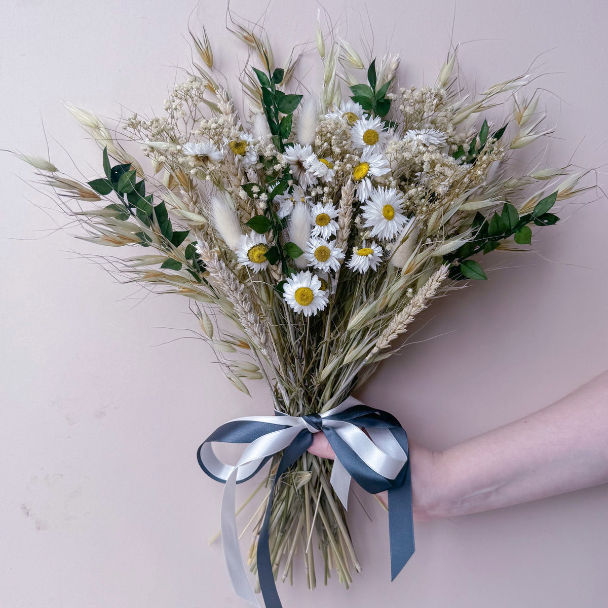 A dried flower bouquet featuring white daisies, wheat and green foliage, held by a person's hand against a soft pink background.