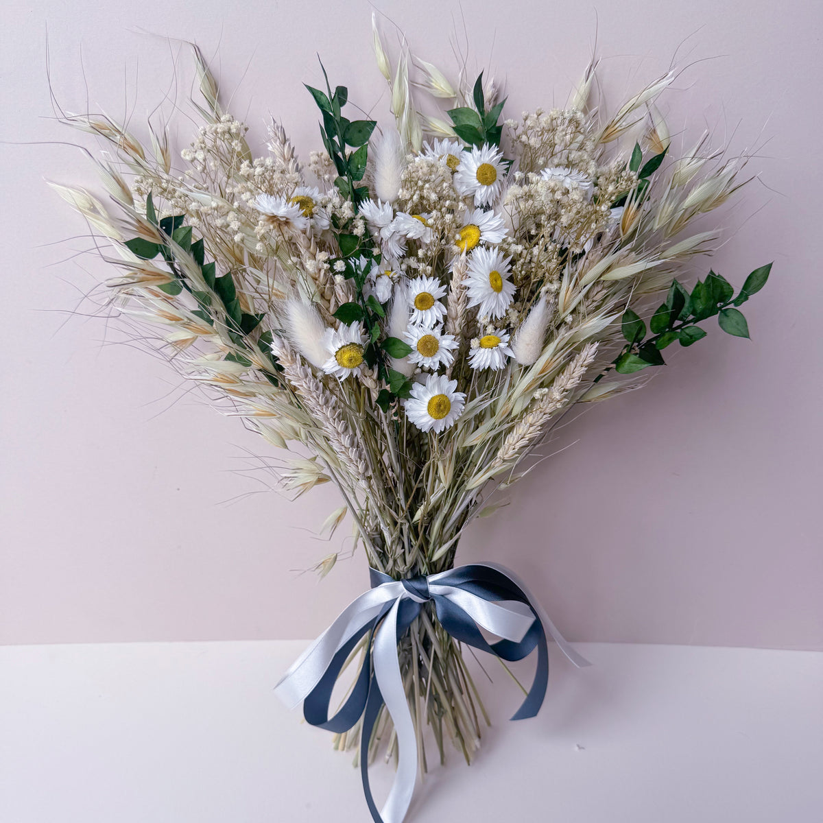 A dried flower bouquet featuring white daisies with yellow centers, natural wheat stalks, and green foliage, tied with a green and white ribbon, set against a soft pink background.