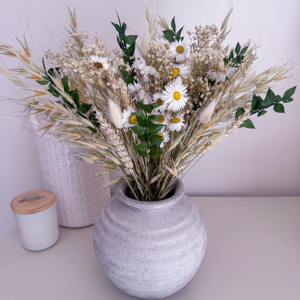 A decorative dried flower arrangement in a textured gray ceramic vase, displayed on a light tabletop with a white candle and a patterned vase in the background.