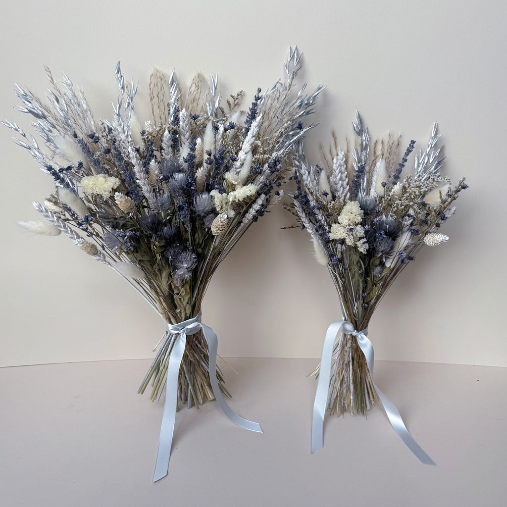 Two dried flower bouquets made from silver-grey, lavender, beige, and white botanicals, tied with pale grey ribbons, set against a plain beige background.