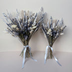 Two dried flower bouquets made from silver-grey, lavender, beige, and white botanicals, tied with pale grey ribbons, set against a plain beige background.