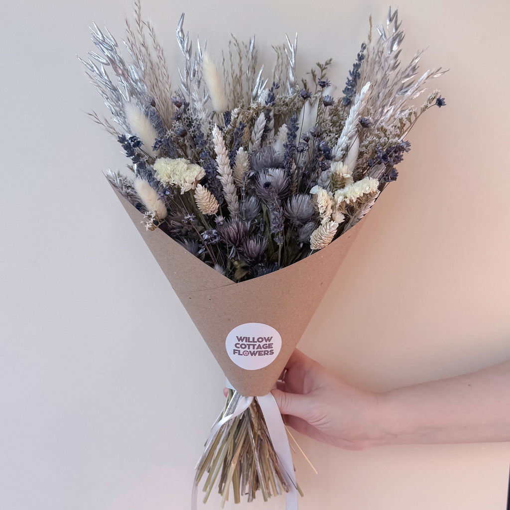 A hand holding a cone-wrapped bouquet of dried flowers and grasses in muted beige, gray, and lavender tones. A round white sticker on the wrap reads 'WILLOW COTTAGE FLOWERS'.