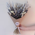 A hand holding a cone-wrapped bouquet of dried flowers and grasses in muted beige, gray, and lavender tones. A round white sticker on the wrap reads 'WILLOW COTTAGE FLOWERS'.