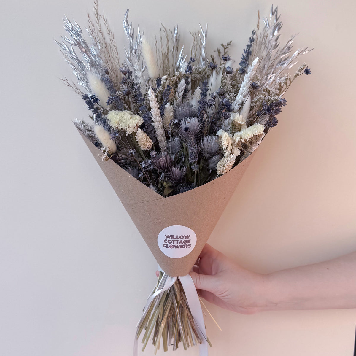 A hand holding a cone-wrapped bouquet of dried flowers and grasses in muted beige, gray, and lavender tones. A round white sticker on the wrap reads 'WILLOW COTTAGE FLOWERS'.