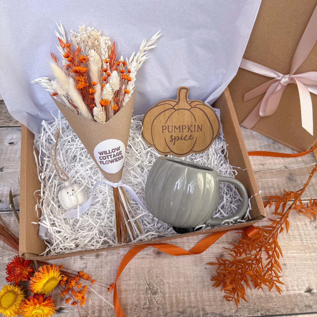 A gift box on a wood surface featuring a dried flower bouquet in a kraft cone, a small white pumpkin ornament, a grey ceramic mug, a wooden pumpkin sign reading 'Pumpkin Spice,' and a kraft gift box with a pink ribbon in the background.