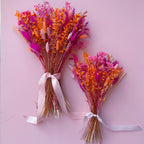 Two dried flower bouquets in pink and orange tones, tied with light pink satin ribbons, arranged on a soft pink background.