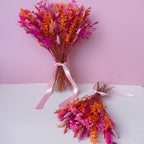 Two small dried flower bouquets in vibrant shades of pink, orange, and cerise, tied with light pink ribbons, displayed on a white surface against a solid pink background.