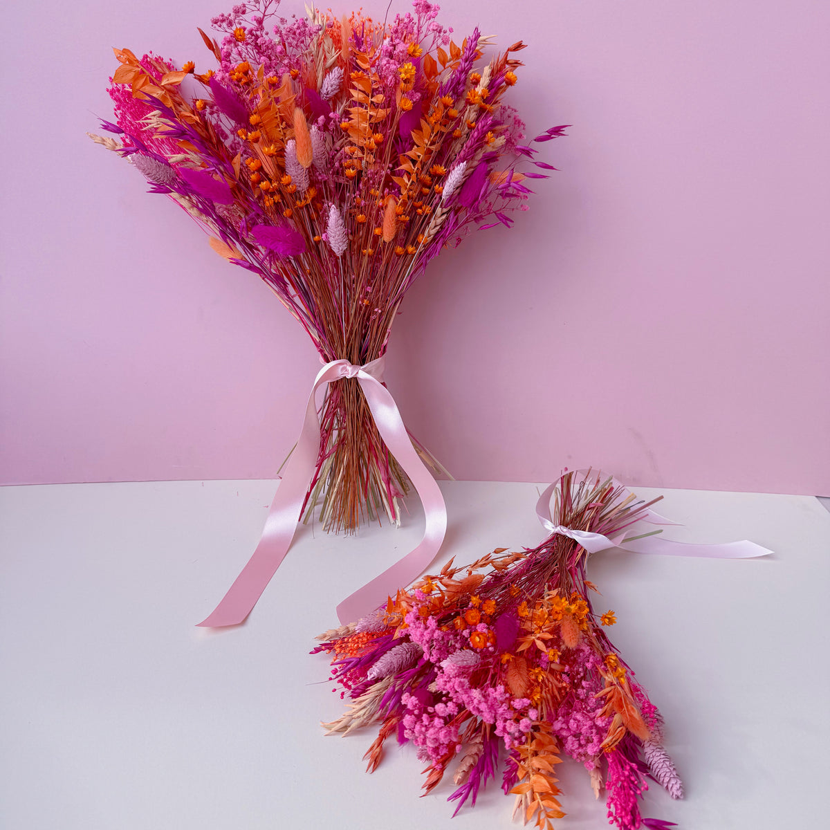 Two small dried flower bouquets in vibrant shades of pink, orange, and cerise, tied with light pink ribbons, displayed on a white surface against a solid pink background.