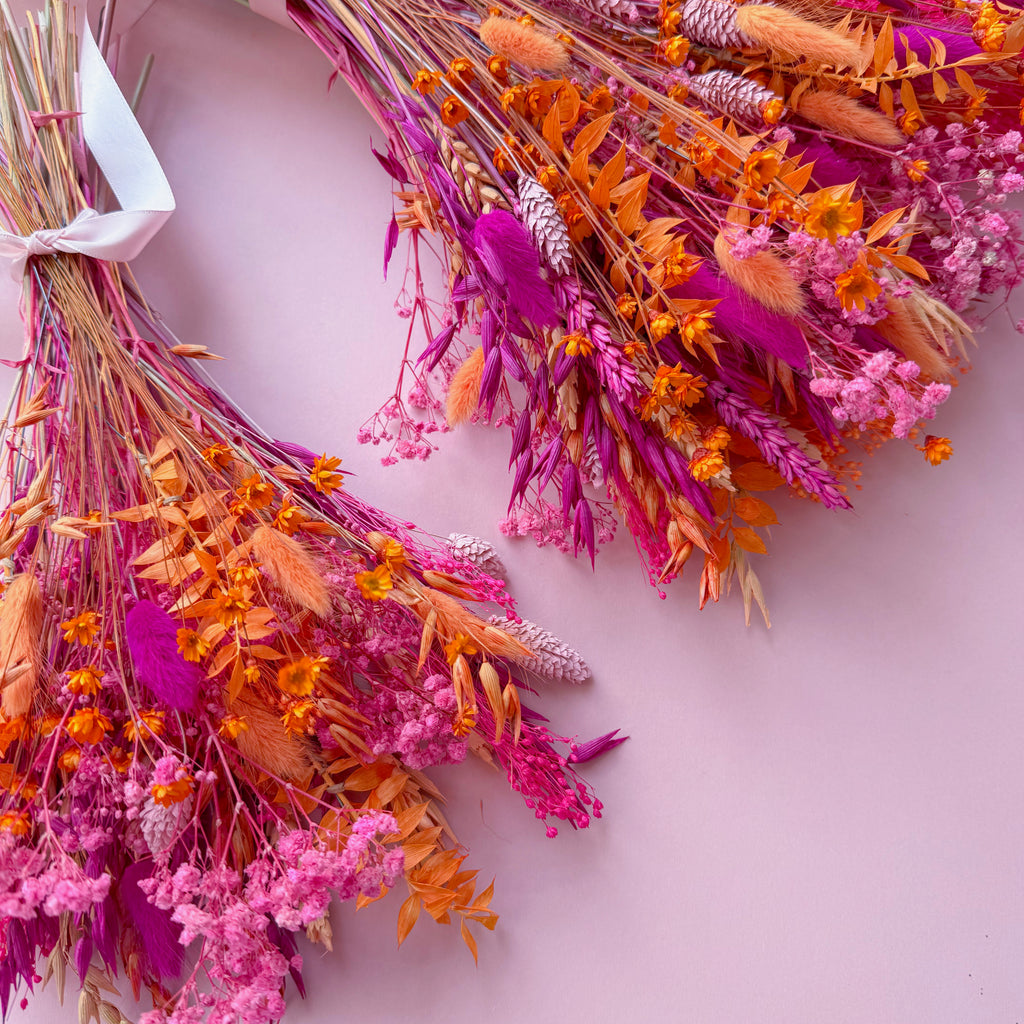 Two round dried flower bouquets in vibrant shades of pink, orange, and cerise, decorated with various grasses, wildflowers, and a light pink ribbon, arranged on a light pink background.