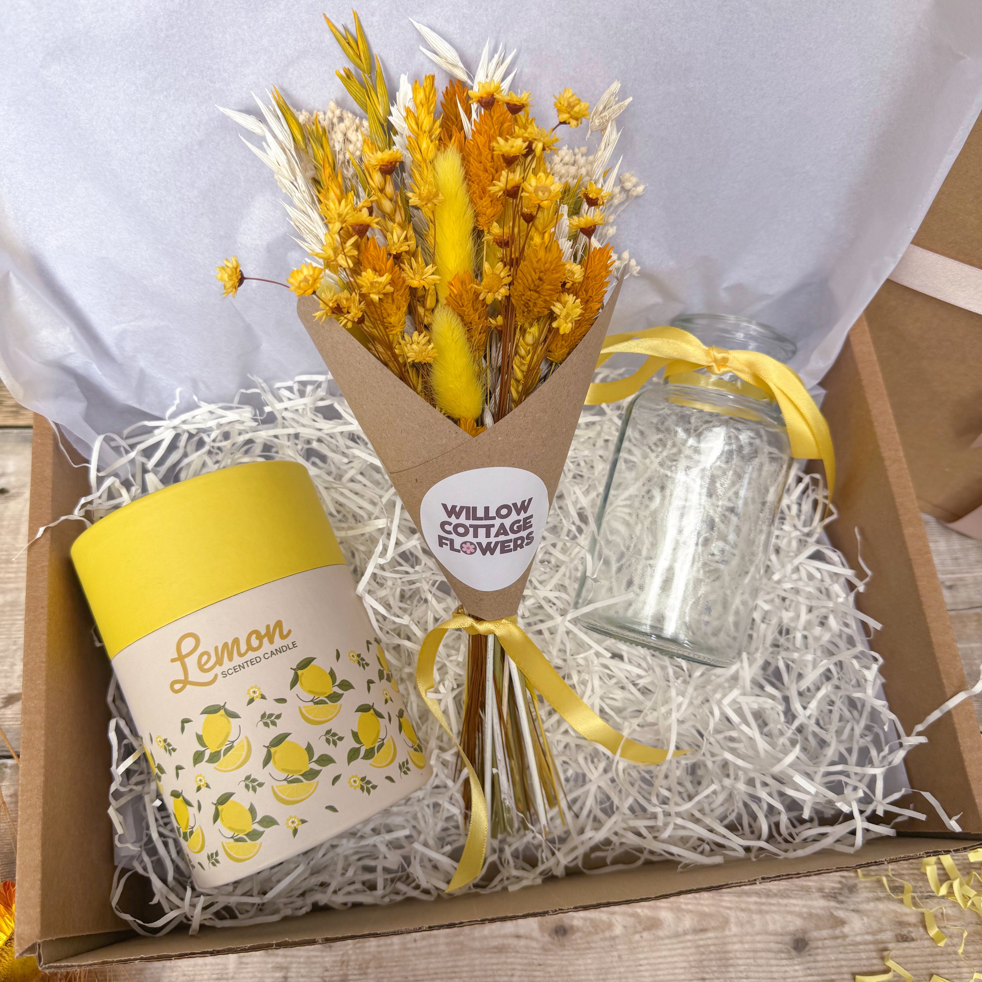 A gift box arrangement featuring a small bouquet of yellow and orange dried flowers wrapped in brown paper, a yellow-lidded lemon-themed tin, and a clear glass jar tied with a yellow ribbon, all set in white shredded paper inside a cardboard box.