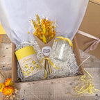 A curated gift box set containing a yellow lemon scented candle, a small yellow dried flower bouquet, and a clear glass vase, arranged in white shredded paper inside a brown cardboard box.