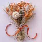 A dried flower bouquet featuring orange bunny tails, light orange baby's breath, a large cream peony, and natural beige grasses tied with an orange ribbon, photographed from above on a light pink background.