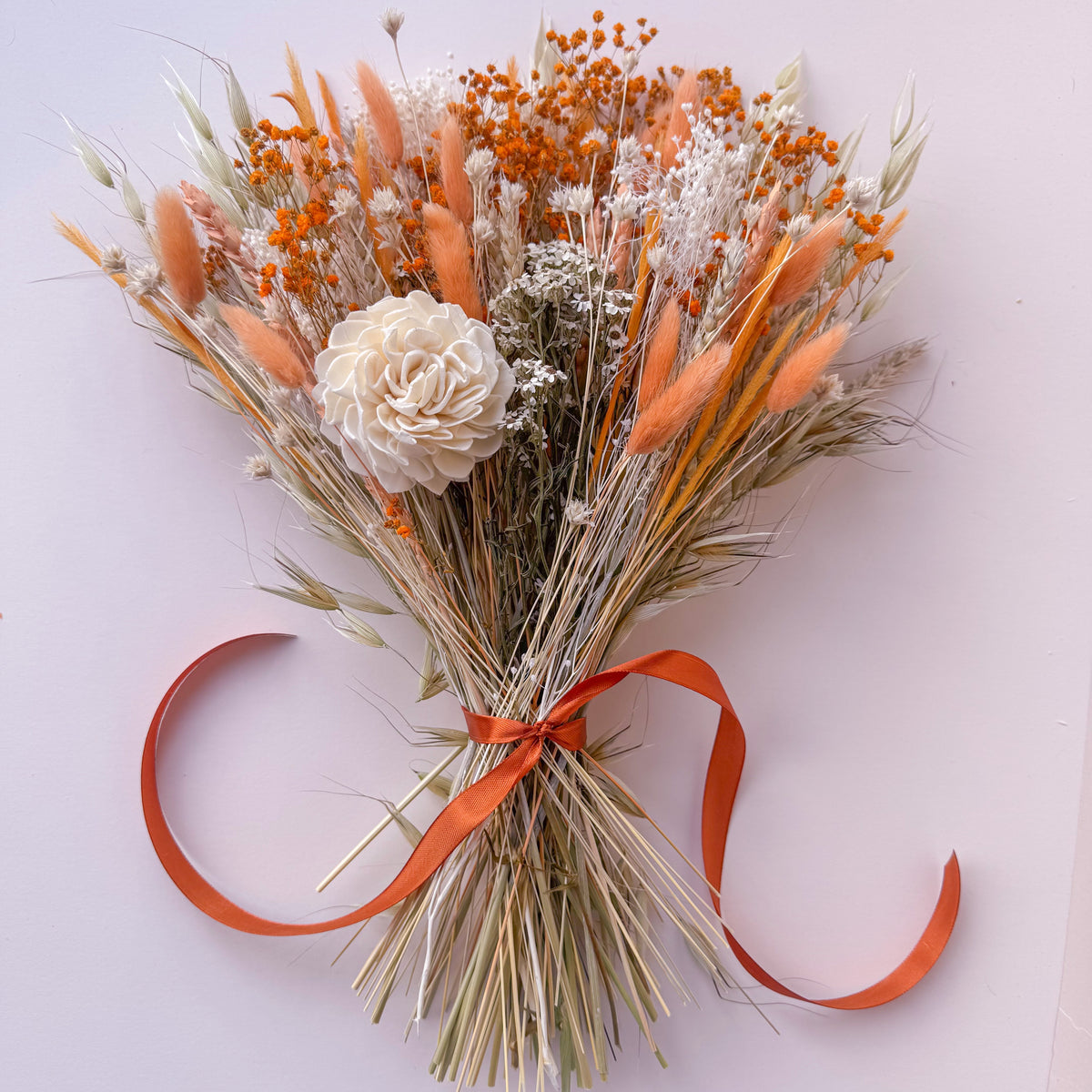 A dried flower bouquet featuring orange bunny tails, light orange baby's breath, a large cream peony, and natural beige grasses tied with an orange ribbon, photographed from above on a light pink background.