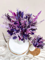 A dried flower arrangement in a white vase with a round white pedestal base, styled on a draped white fabric surface with neutral decor. The bouquet features a mix of purple, violet, lavender, and pink dried flowers and grasses.