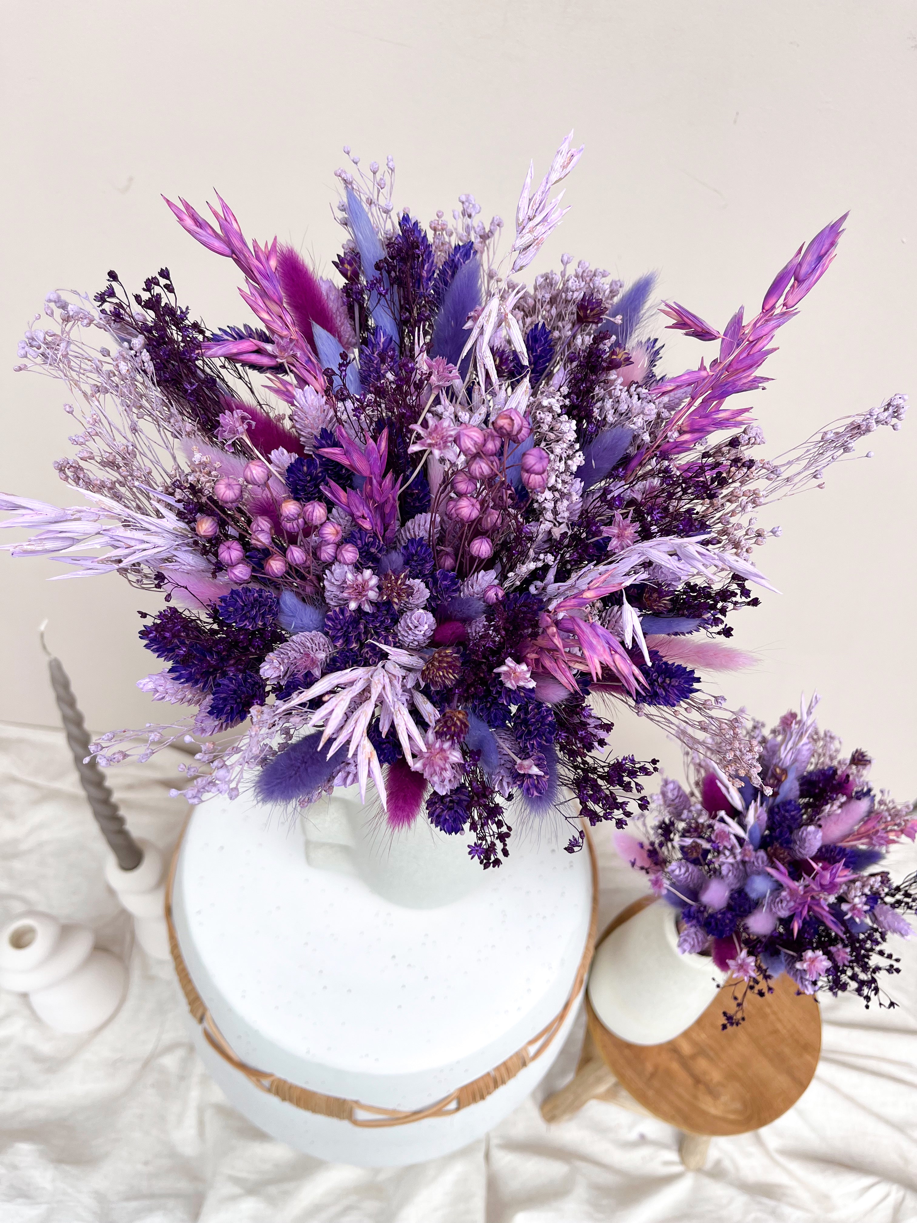 A dried flower arrangement in a white vase with a round white pedestal base, styled on a draped white fabric surface with neutral decor. The bouquet features a mix of purple, violet, lavender, and pink dried flowers and grasses.