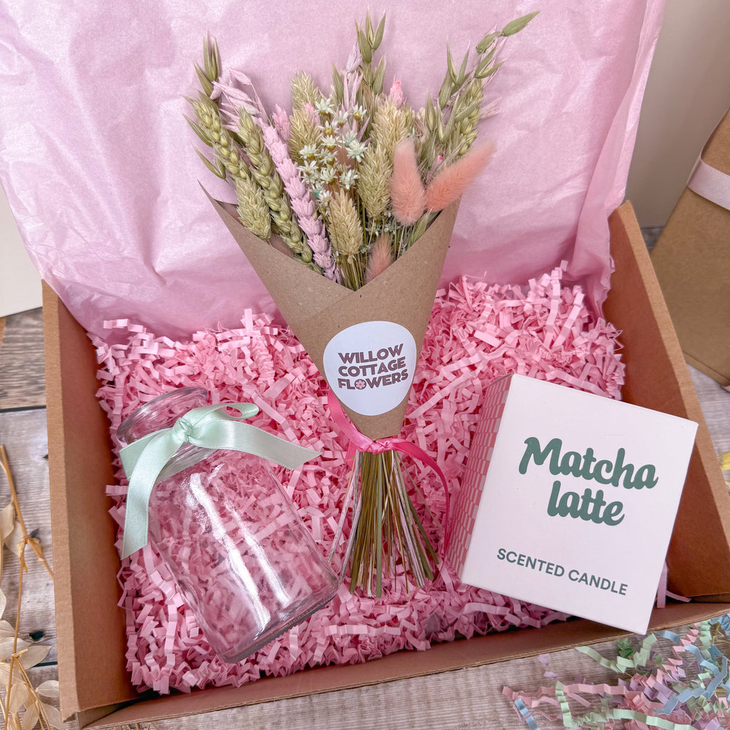 A gift box arrangement featuring a small bouquet of dried flowers in a brown paper wrap, a clear glass jar tied with a mint green ribbon and a 'Matcha latte SCENTED CANDLE,' all set in pink shredded paper inside a cardboard box.