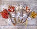 Five small dried flower bouquets wrapped in brown kraft paper, arranged on a rustic grey wooden surface. The bouquets contain mixed dried flowers in red, orange, cream, yellow, and gold tones with metallic gold and white ribbons tied at the base.