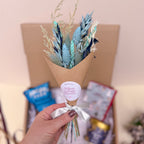 Blue dried flower Bouquet in gift wrap with 'Willow Cottage Flowers' label held by a hand against a blurred background.