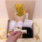 Bouquet of yellow flowers wrapped in brown paper with a white ribbon held by a hand, against a blurred indoor background.