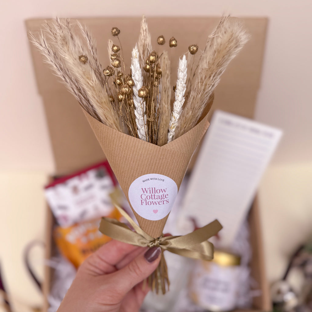 Bouquet of dried flowers in a brown paper cone with a gold ribbon, held by a hand.