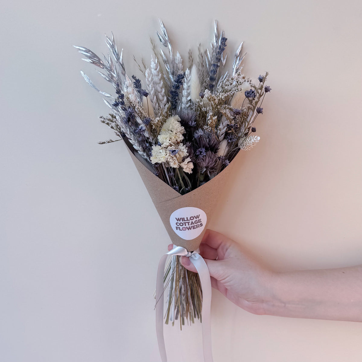 A hand holding a small bouquet of dried flowers wrapped in brown kraft paper with a white round sticker that reads 'WILLOW COTTAGE FLOWERS'. The bouquet contains silver-toned grasses, lavender, and cream blossoms against a plain light beige background.