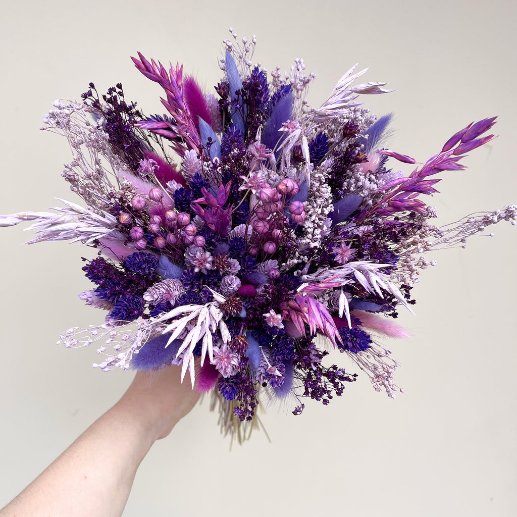 A hand holding a bridesmaid dried flower bouquet in shades of purple, violet, and pink against a plain light beige background.