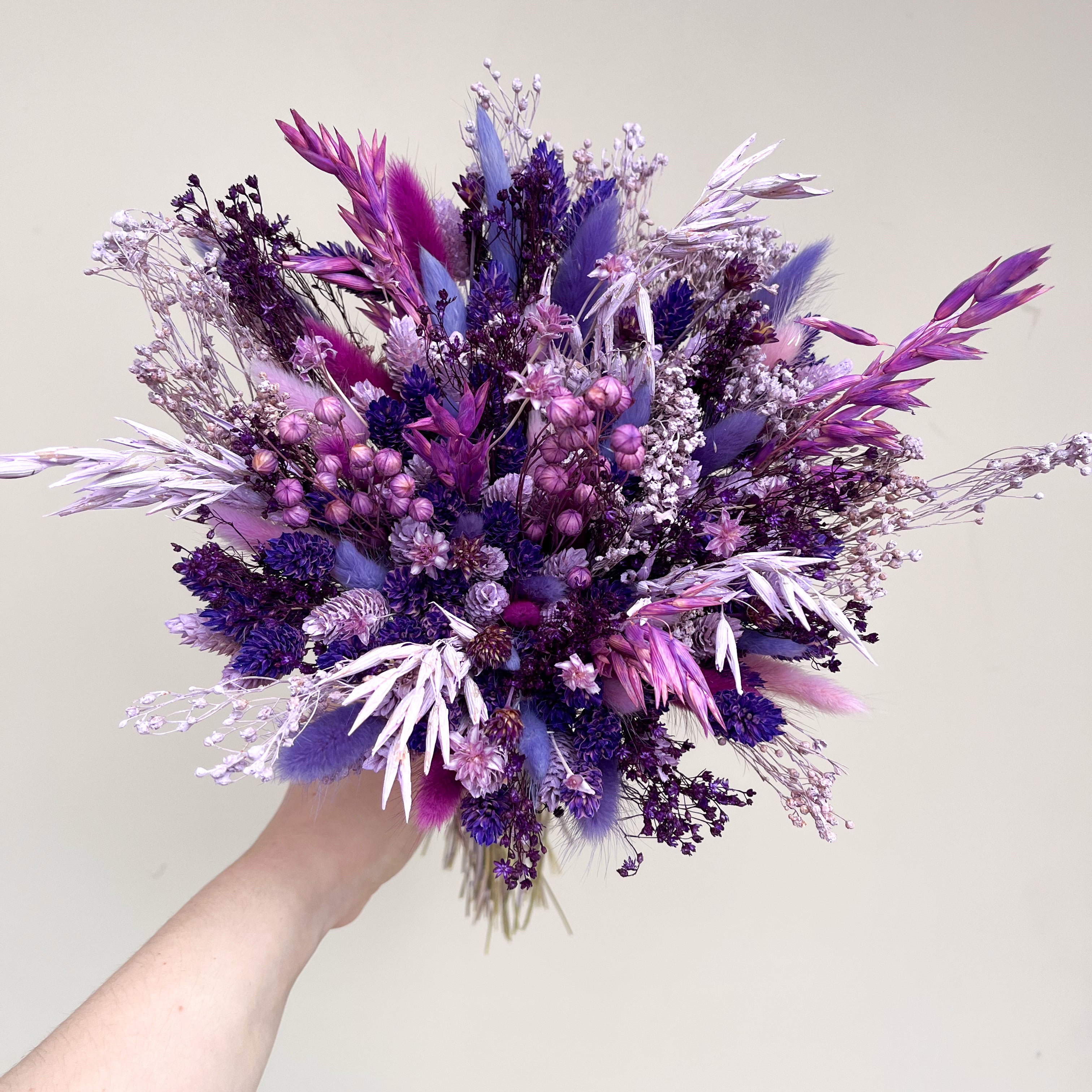 A hand holding a bridesmaid dried flower bouquet in shades of purple, violet, and pink against a plain light beige background.
