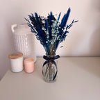 A dried flower bouquet in dark blue in a clear glass vase with a dark navy ribbon, placed on a light beige tabletop next to a white textured ceramic jar and a pink candle.