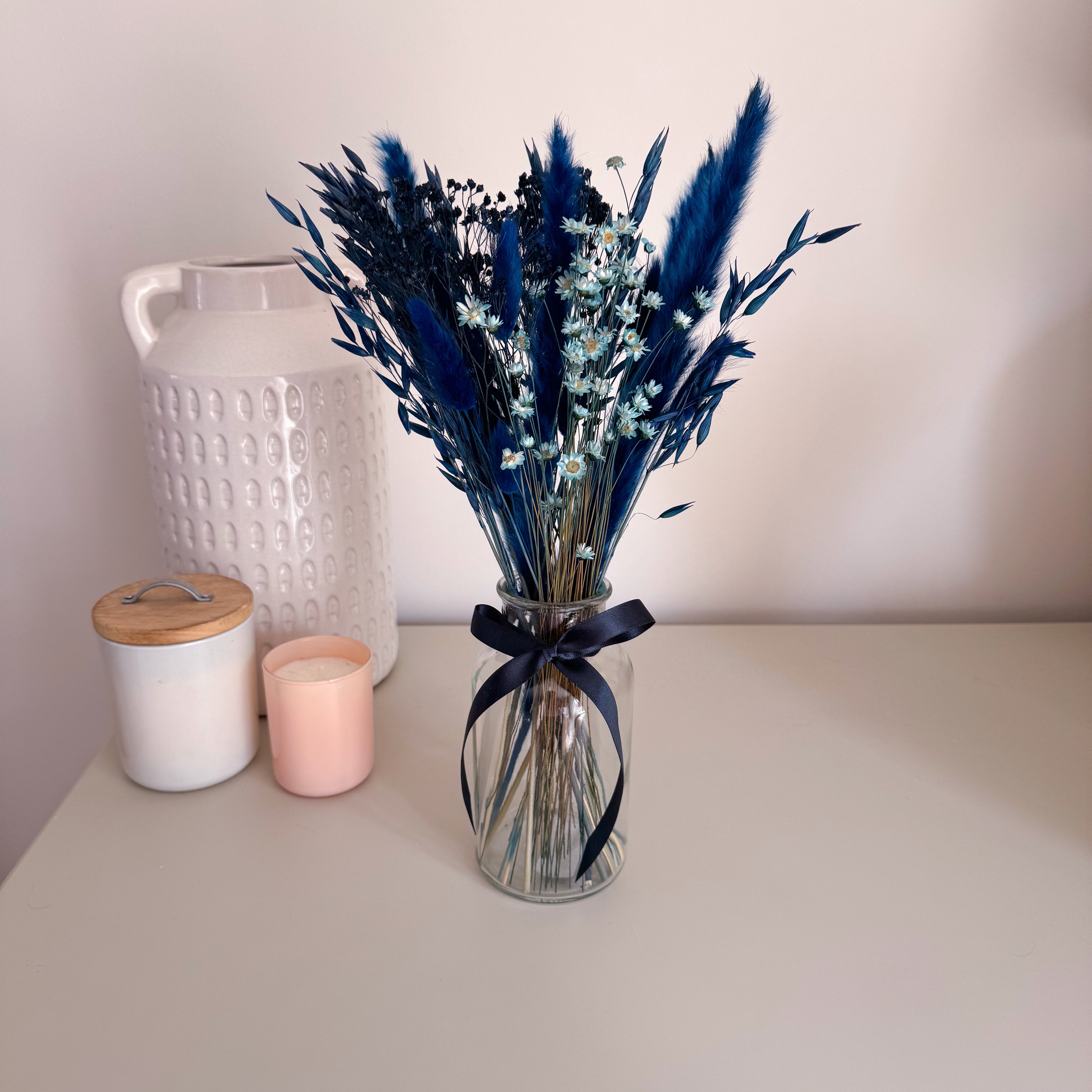 A dried flower bouquet in dark blue in a clear glass vase with a dark navy ribbon, placed on a light beige tabletop next to a white textured ceramic jar and a pink candle.