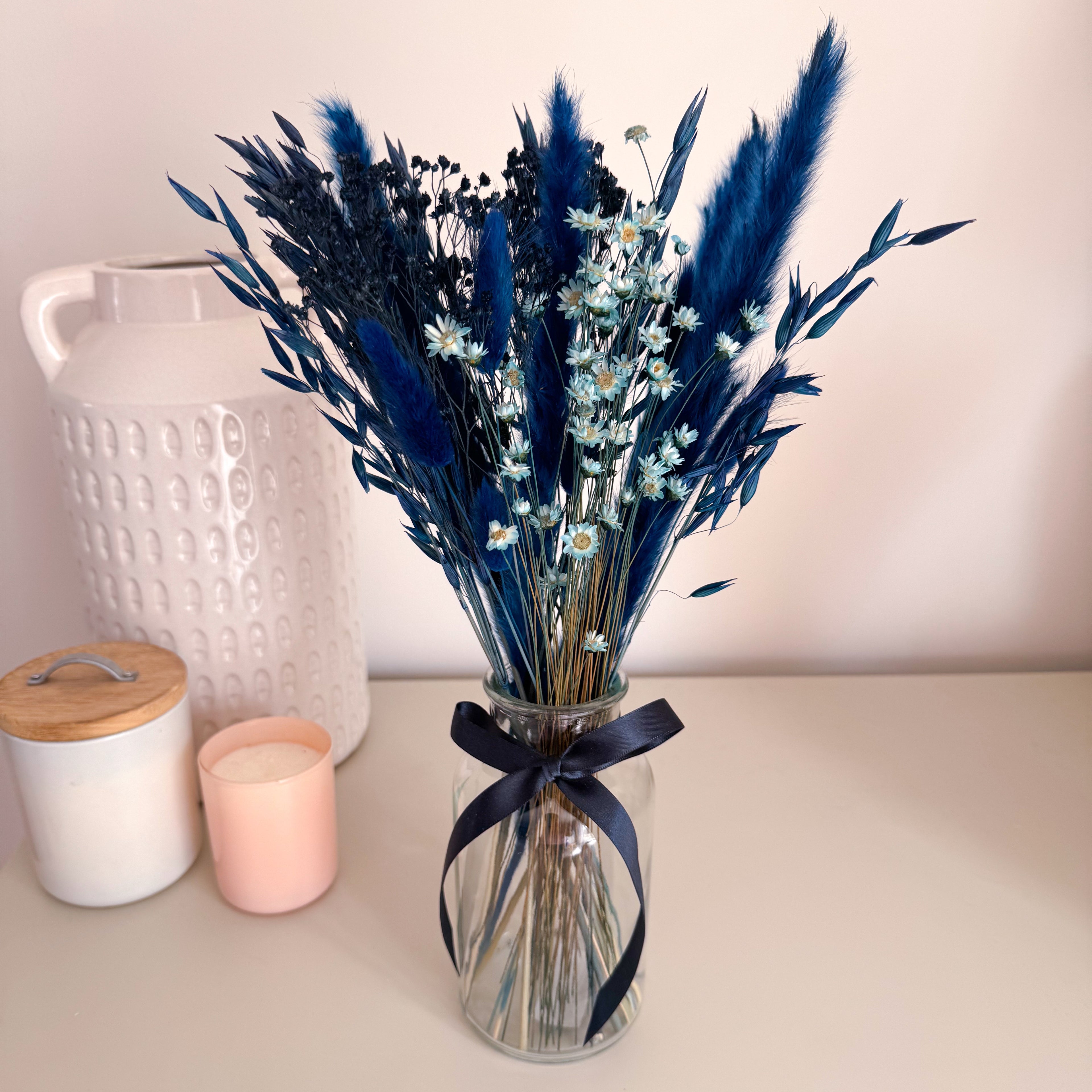 A dried navy blue pampas grass arrangement in a clear glass vase with a navy ribbon, displayed on a tabletop beside a white textured vase and a pink candle.
