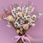 A dried flower bouquet held by a hand against a solid pink background. The bouquet features white daisies, pale pink and cream strawflowers, pink filler flowers, and fluffy white bunny tails tied with a light pink ribbon.