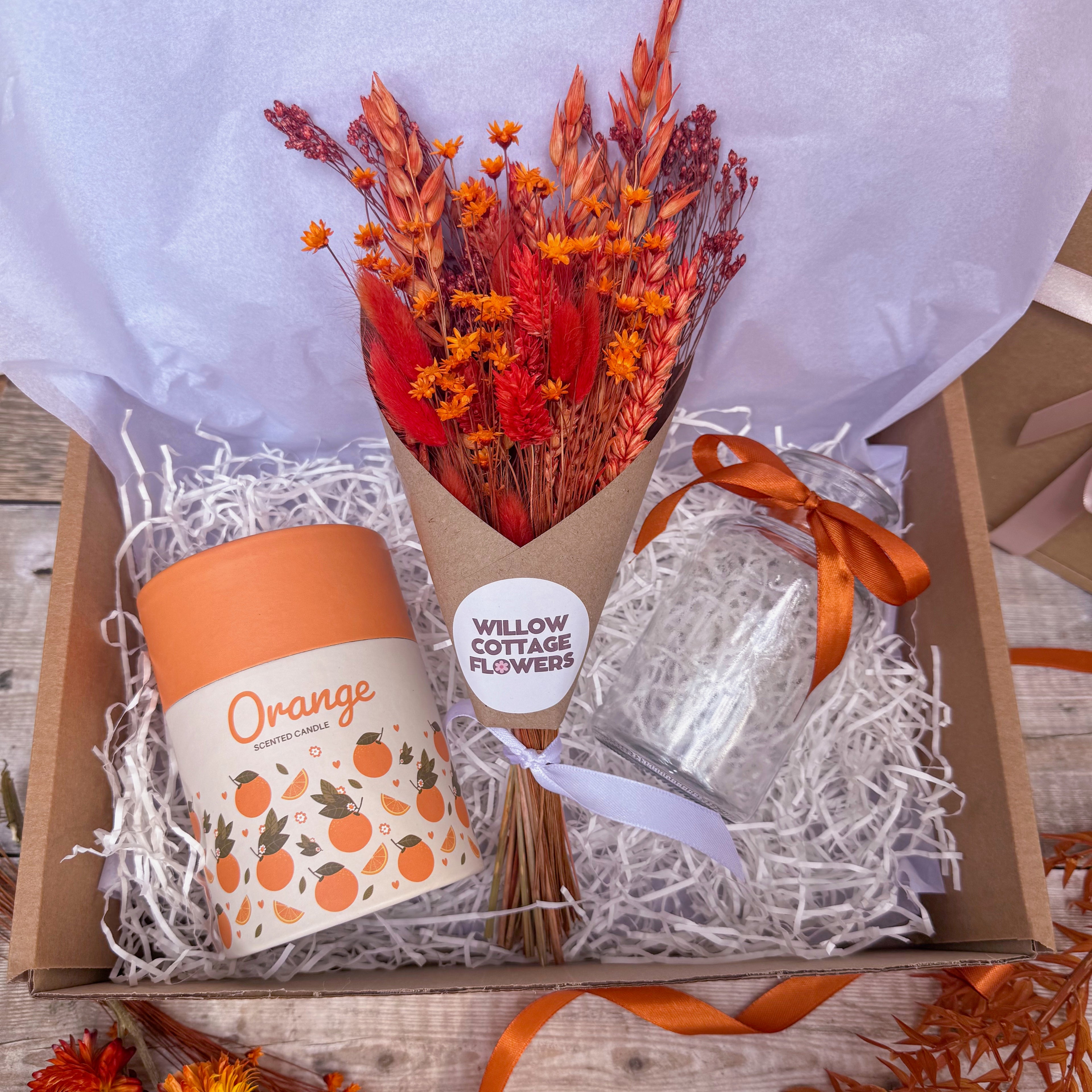 A gift box arrangement featuring a dried flower bouquet with orange florals in a brown paper wrap, an orange scented candle and a clear glass vase tied with an orange ribbon, all nestled in white paper shred inside a cardboard box.