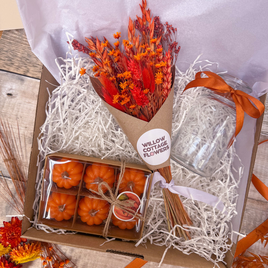 An autumn themed gift box from Willow Cottage Flowers, filled with white shredded paper. Inside, there is a small bouquet of orange dried flowers in a brown kraft paper cone, a clear glass vase with an orange ribbon and a boxed set of six mini orange pumpkin candles.