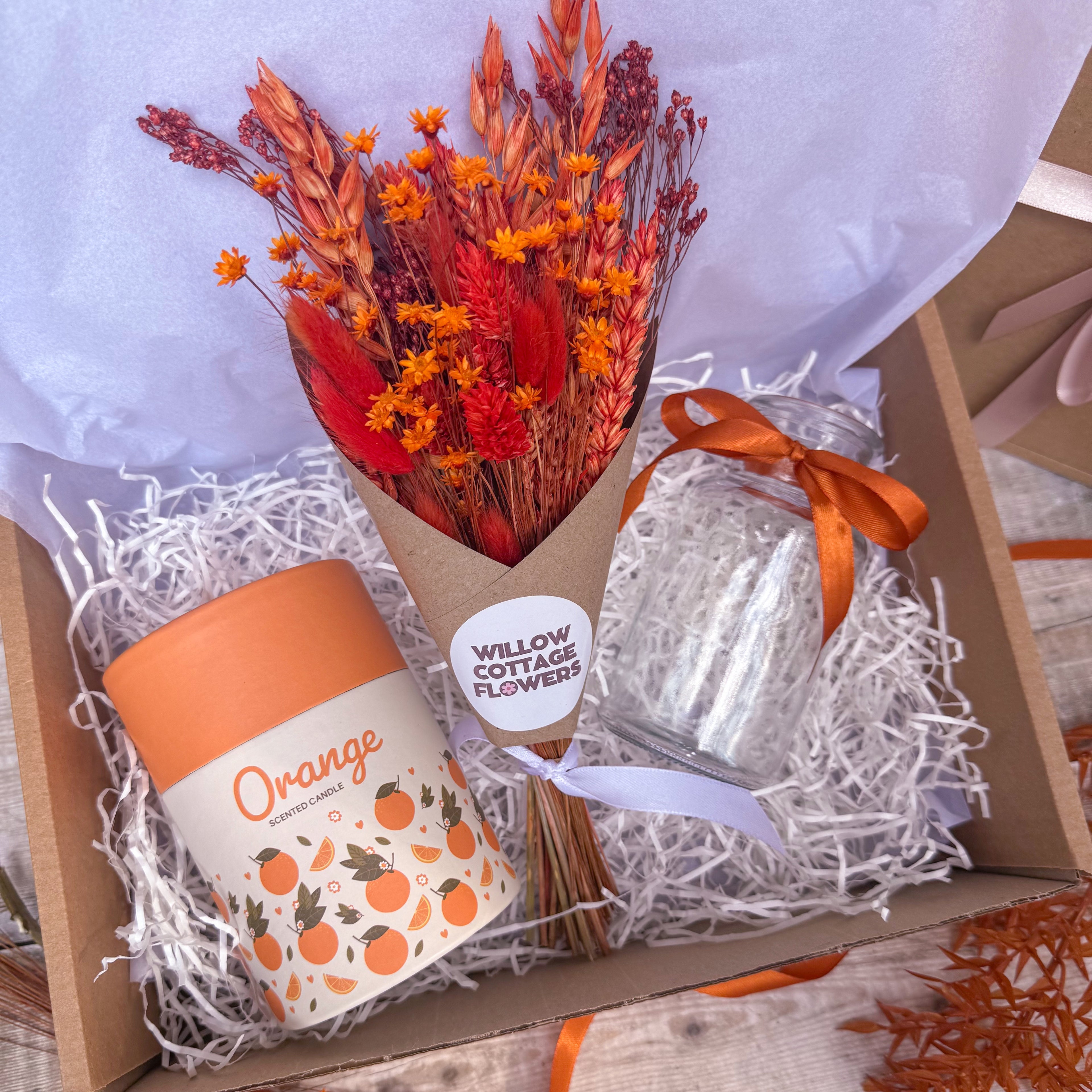 A gift box arrangement featuring a dried flower bouquet with orange wrapped in kraft paper, an orange scented candle and a clear glass vase tied with an orange ribbon, all nestled in white shredded paper.