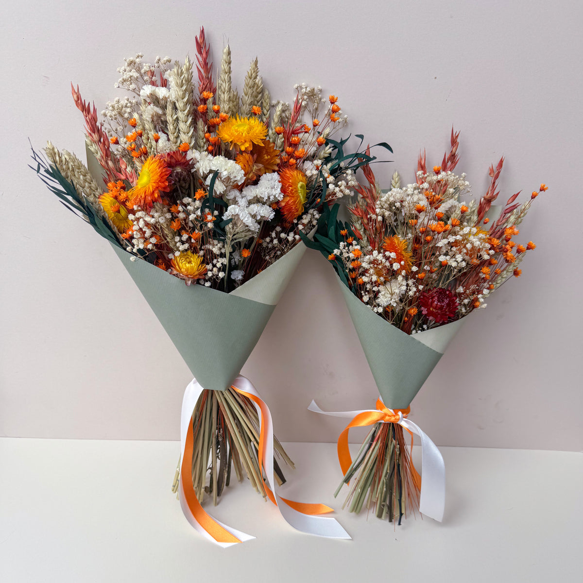 Two dried flower bouquets wrapped in sage green paper with orange and white ribbons, displayed on a white surface against a light grey background