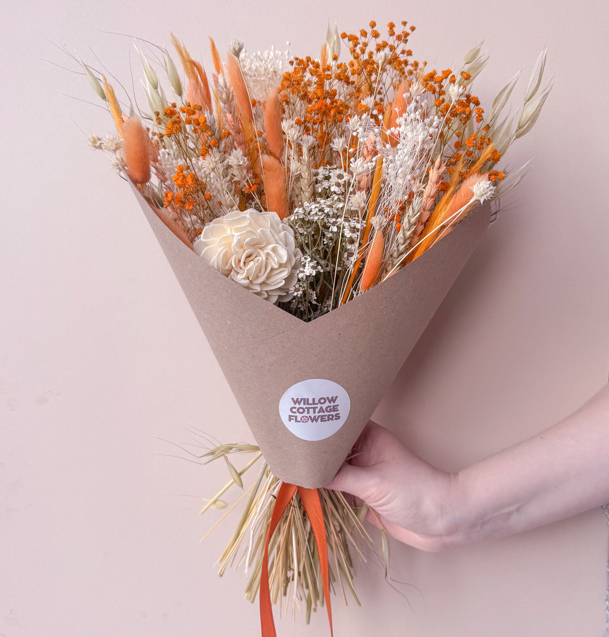A hand holding an orange dried flower bouquet wrapped in brown kraft paper with a white circular 'WILLOW COTTAGE FLOWERS' sticker. The bouquet features orange wheat, baby's breath, and a cream-coloured sola flower against a solid pale pink background.