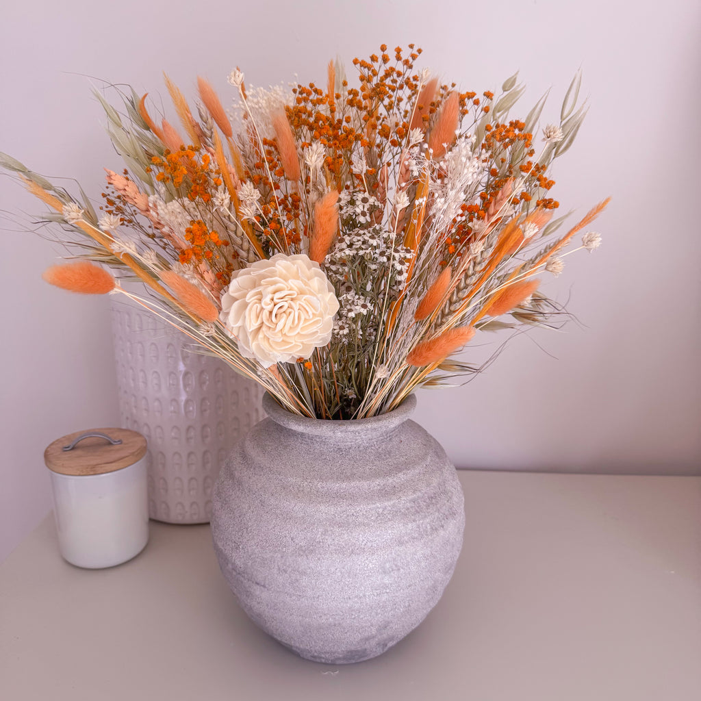 A ceramic grey vase filled with a dried floral arrangement featuring orange reeds, white baby's breath, and a large cream peony, displayed on a tabletop next to a candle.