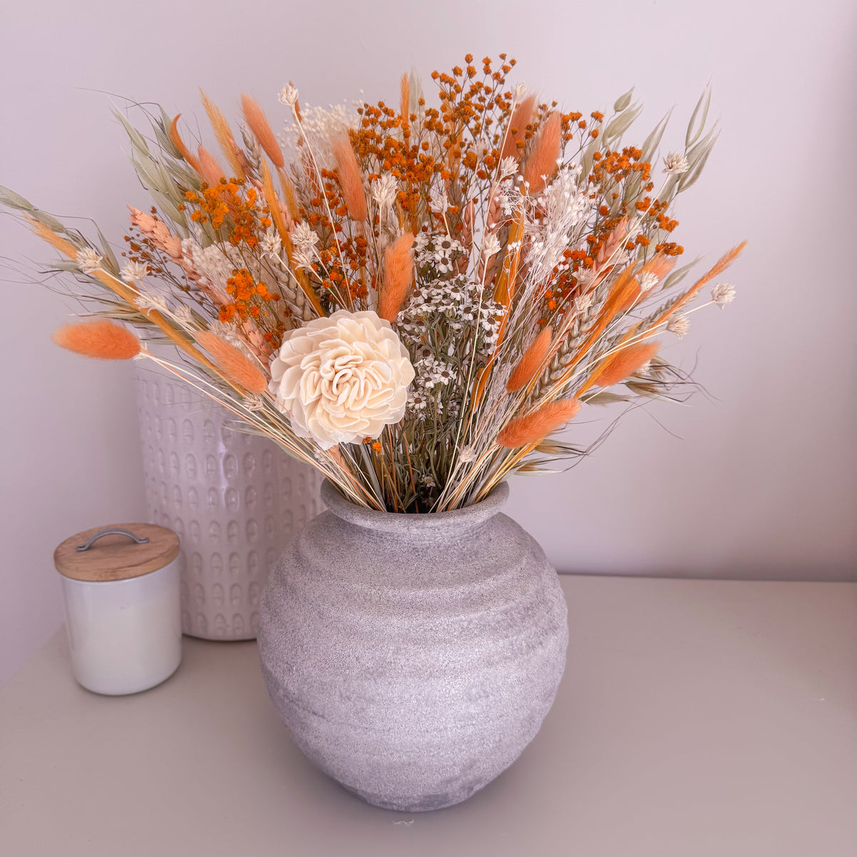 A ceramic grey vase filled with a dried floral arrangement featuring orange reeds, white baby's breath, and a large cream peony, displayed on a tabletop next to a candle.